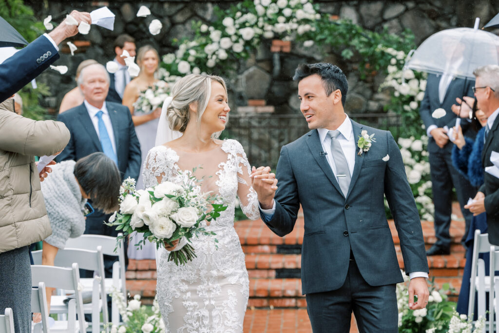 Bride and groom walk down the aisle holding hands, surrounded by guests. The bride holds a bouquet, and guests are throwing flower petals.