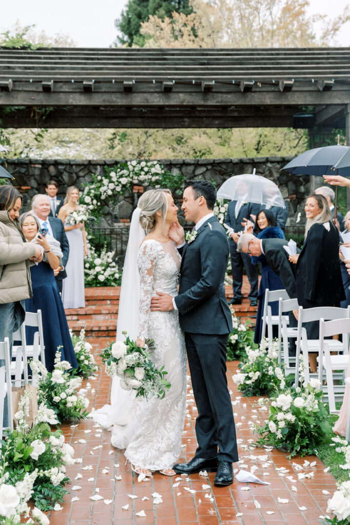 A bride and groom kiss at the end of an outdoor aisle. Guests surround them, some holding umbrellas. White flower arrangements line the aisle.