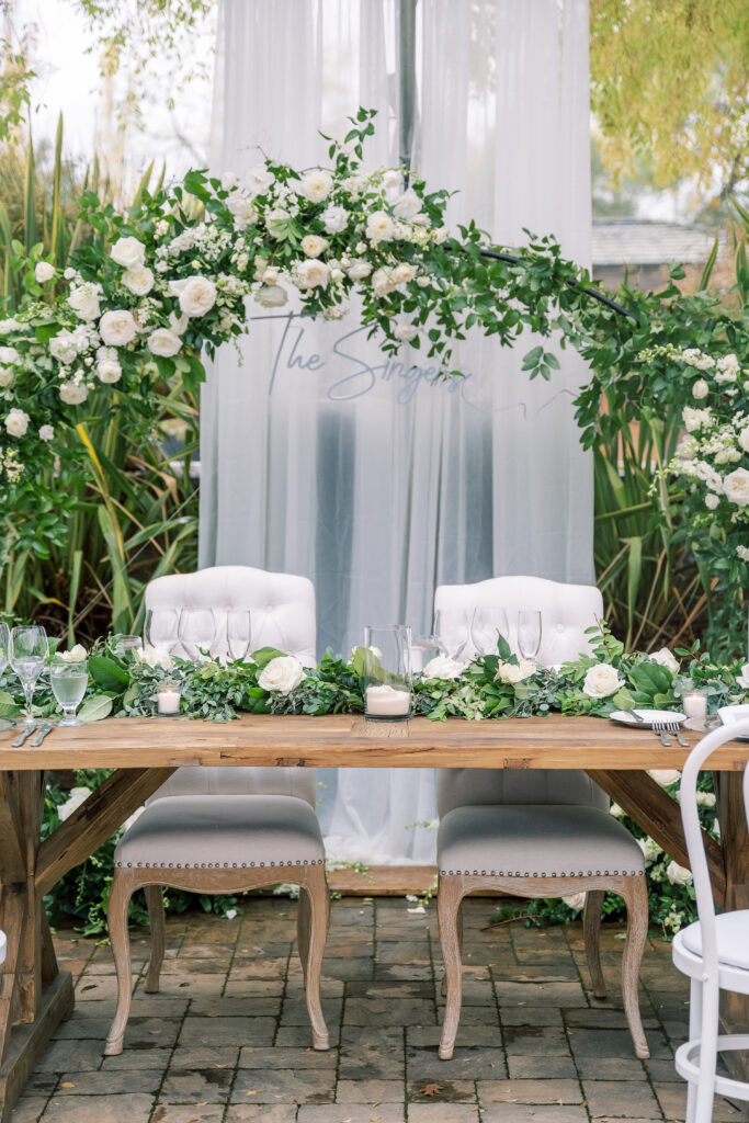 Elegant wedding table with two white chairs, adorned with white flowers and greenery, set under an arch of similar floral arrangement.
