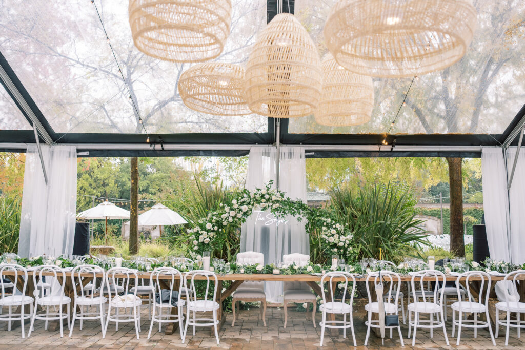 Elegant outdoor wedding reception setup with a long wooden table, white chairs, and green floral arch under a transparent tent. Wicker lights hang from above.