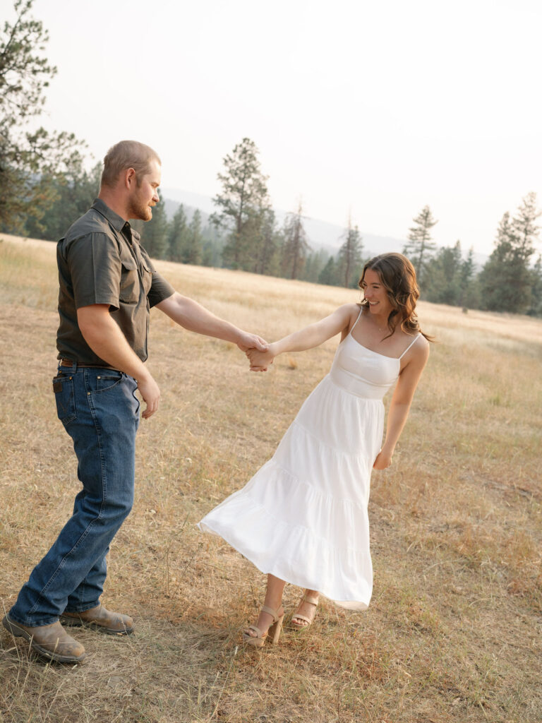 A man and woman hold hands in a field, smiling at each other. The woman wears a white dress, and the man wears a dark shirt and jeans. Trees and hills are visible in the background.