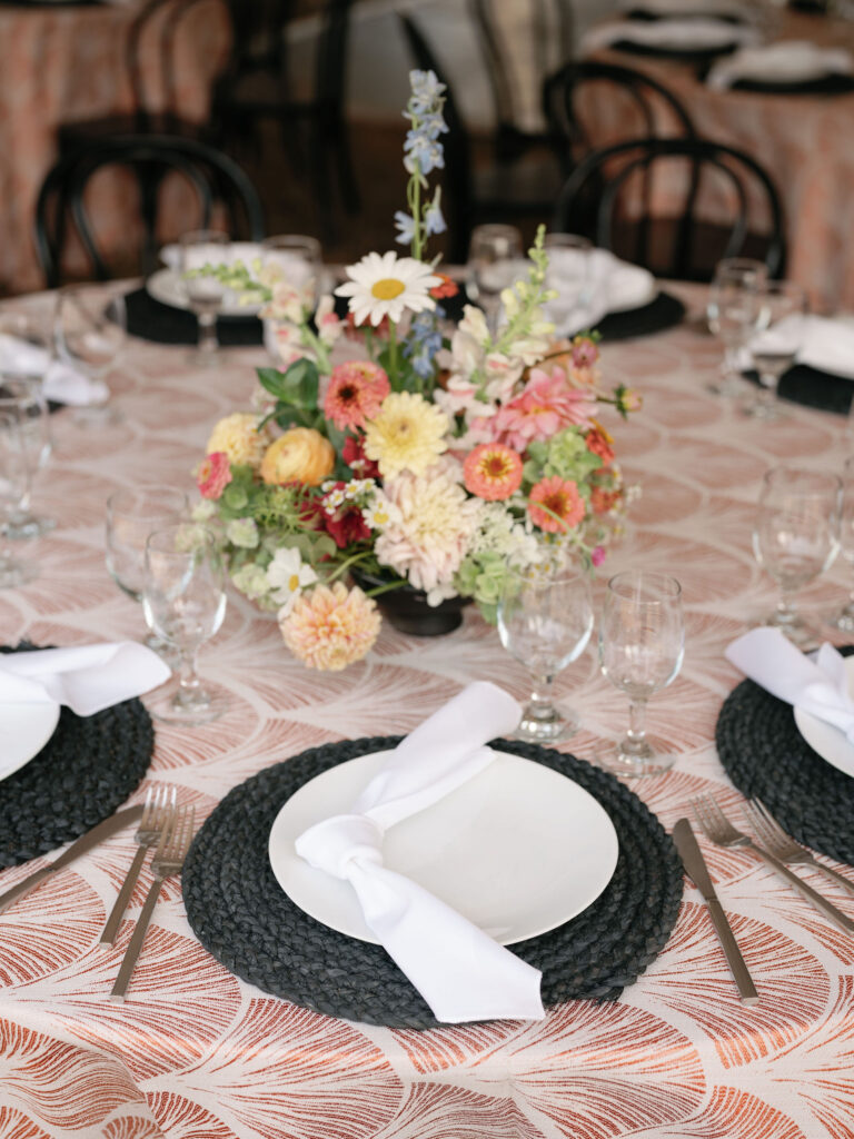A round table set for a meal, featuring a floral centerpiece, patterned tablecloth, black placemats, white plates, white napkins, silver cutlery, and clear glasses.