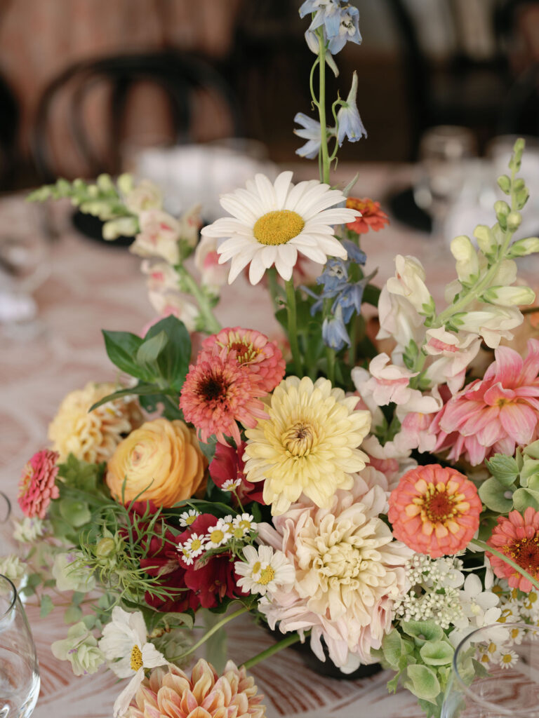 A colorful floral arrangement with daisies, zinnias, snapdragons, and various blossoms on a patterned tablecloth.