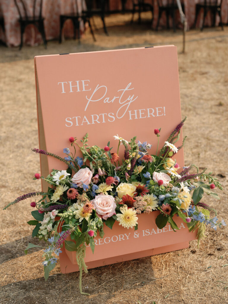 A pink sign with "The Party Starts Here!" and floral arrangements at the bottom. The ground is dry grass.