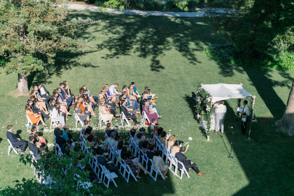 Outdoor wedding ceremony with guests seated on chairs, facing a couple under a floral canopy. The setting is grassy with trees providing shade.