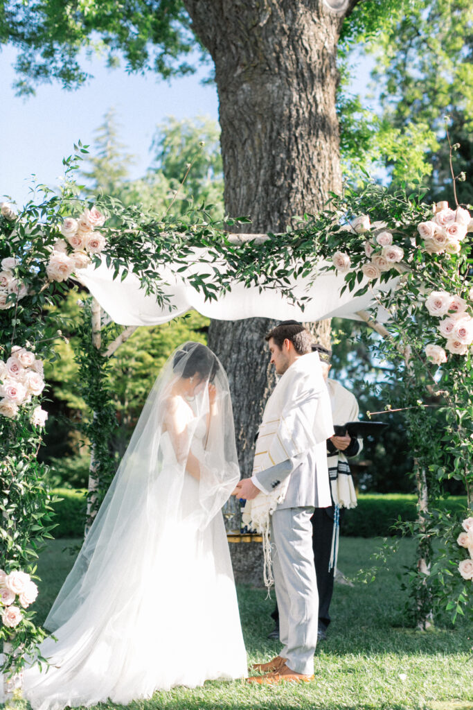 A bride and groom stand under a floral wedding arch outdoors, with a large tree in the background. The groom holds a cloth, and both are dressed in formal attire.