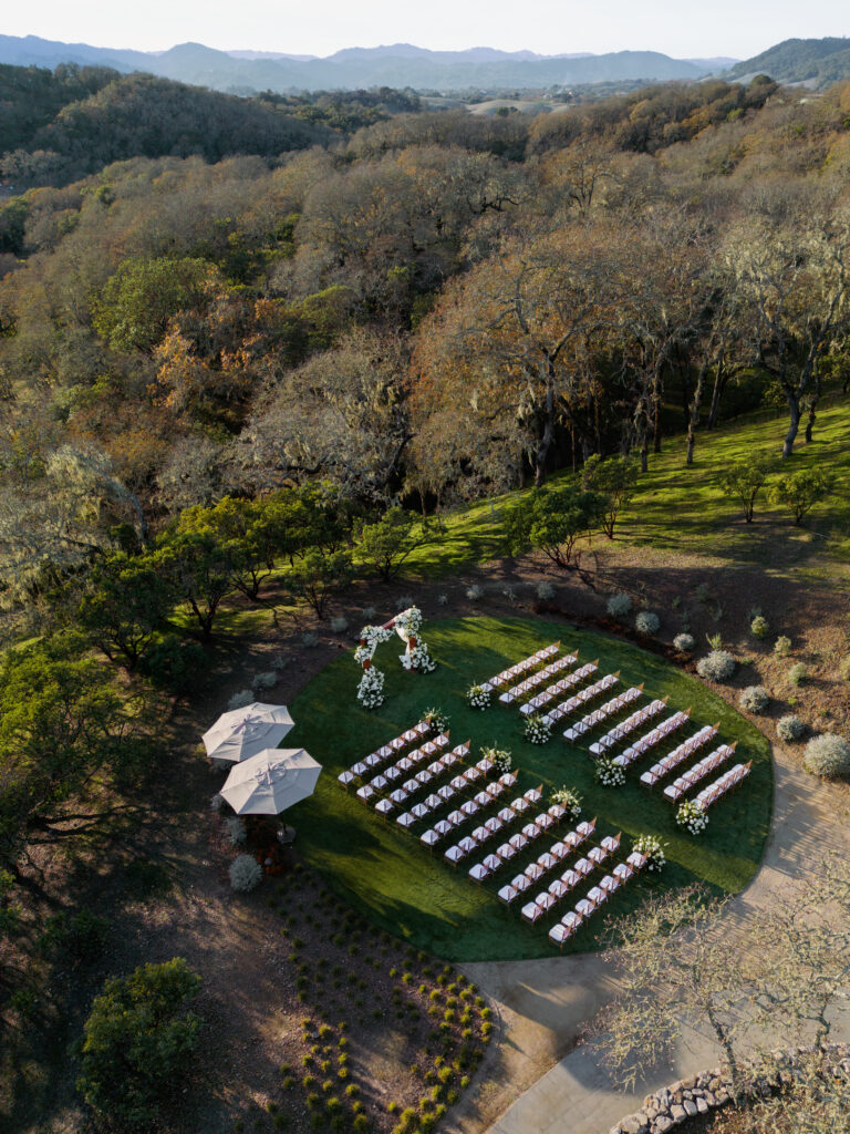 Aerial view of an outdoor wedding setup in a wooded area with rows of white chairs and a floral arch on a circular green lawn, surrounded by trees and hills.