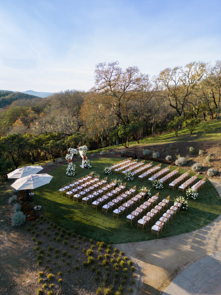 An outdoor wedding setup on grass with rows of chairs facing an arch adorned with flowers, surrounded by trees under a clear sky.