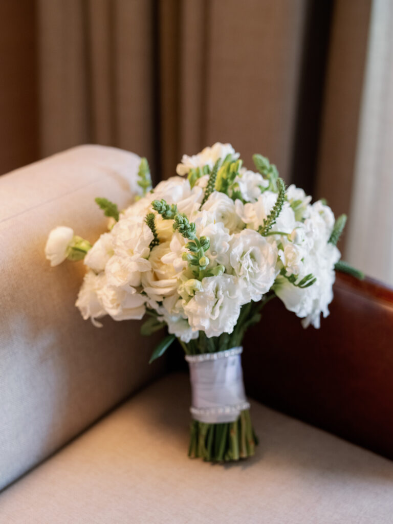 A bouquet of white flowers, including roses and snapdragons, wrapped in white ribbon, rests on a beige sofa.