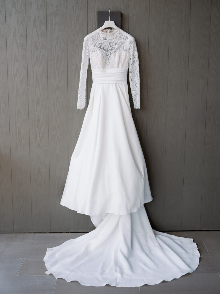 A white wedding dress with lace sleeves and a flowing train hangs against a gray textured wall.
