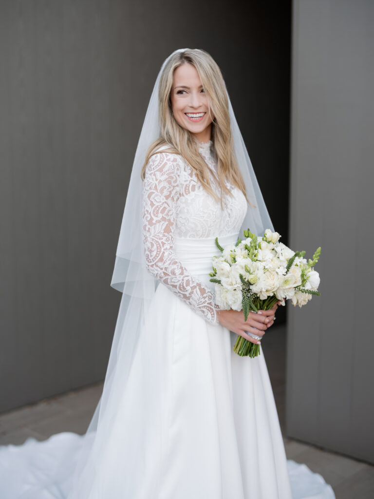 A bride in a white lace dress and veil holds a bouquet of white flowers, smiling.