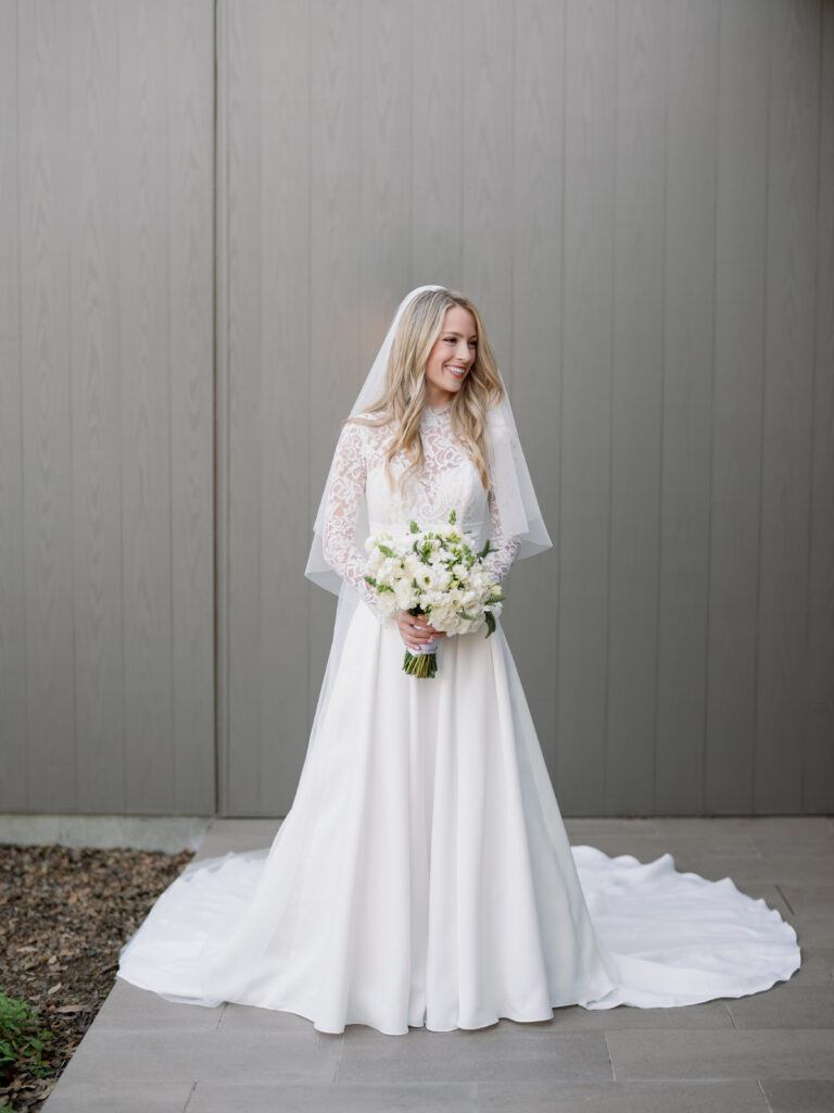 Bride in a white lace gown holding a bouquet of flowers, standing against a gray wall.