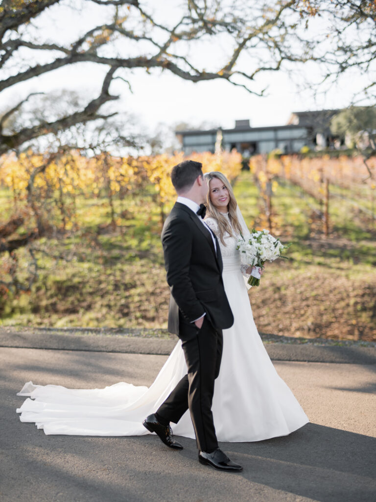 A bride and groom walk along a path with a vineyard in the background.