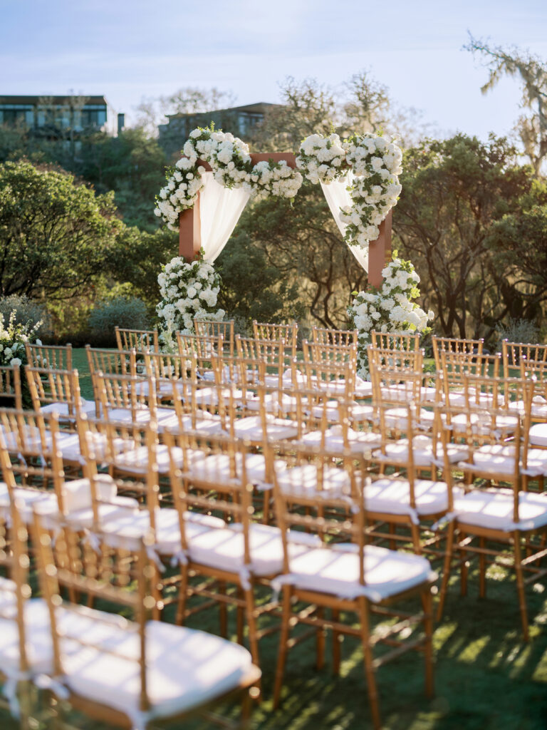 Outdoor wedding setup with a wooden arch adorned with white flowers and draped fabric, surrounded by rows of empty wooden chairs on a grassy area.
