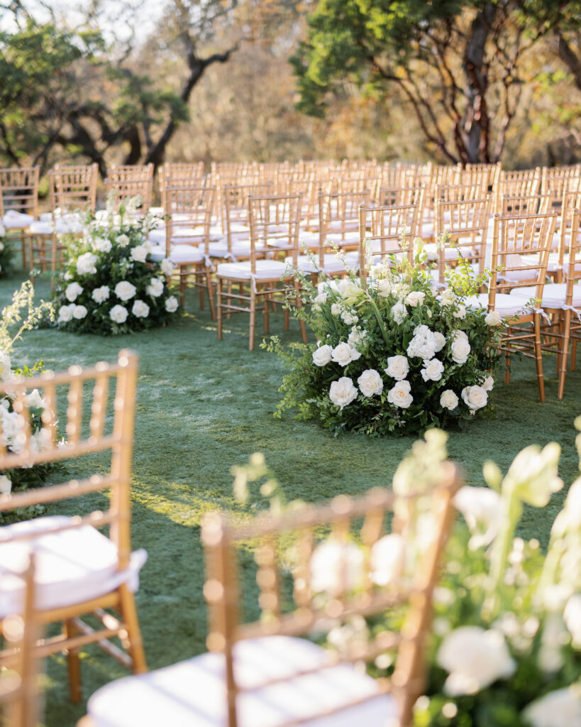 Outdoor wedding ceremony setup with rows of wooden chairs and large floral arrangements of white roses on grassy ground.