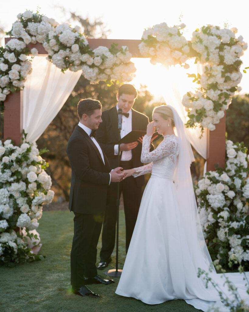 A couple exchanges rings during an outdoor wedding ceremony beneath a flower-adorned arch, with sunlight streaming through, officiated by a man in a tuxedo.