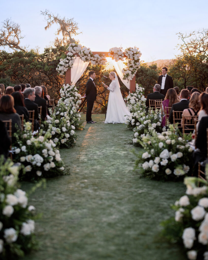 Bride and groom stand under a flower-adorned arch during an outdoor wedding ceremony at sunset, with guests seated on either side of the aisle lined with white floral arrangements.