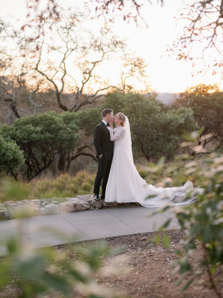A bride and groom share a kiss on a stone path surrounded by trees at sunset.
