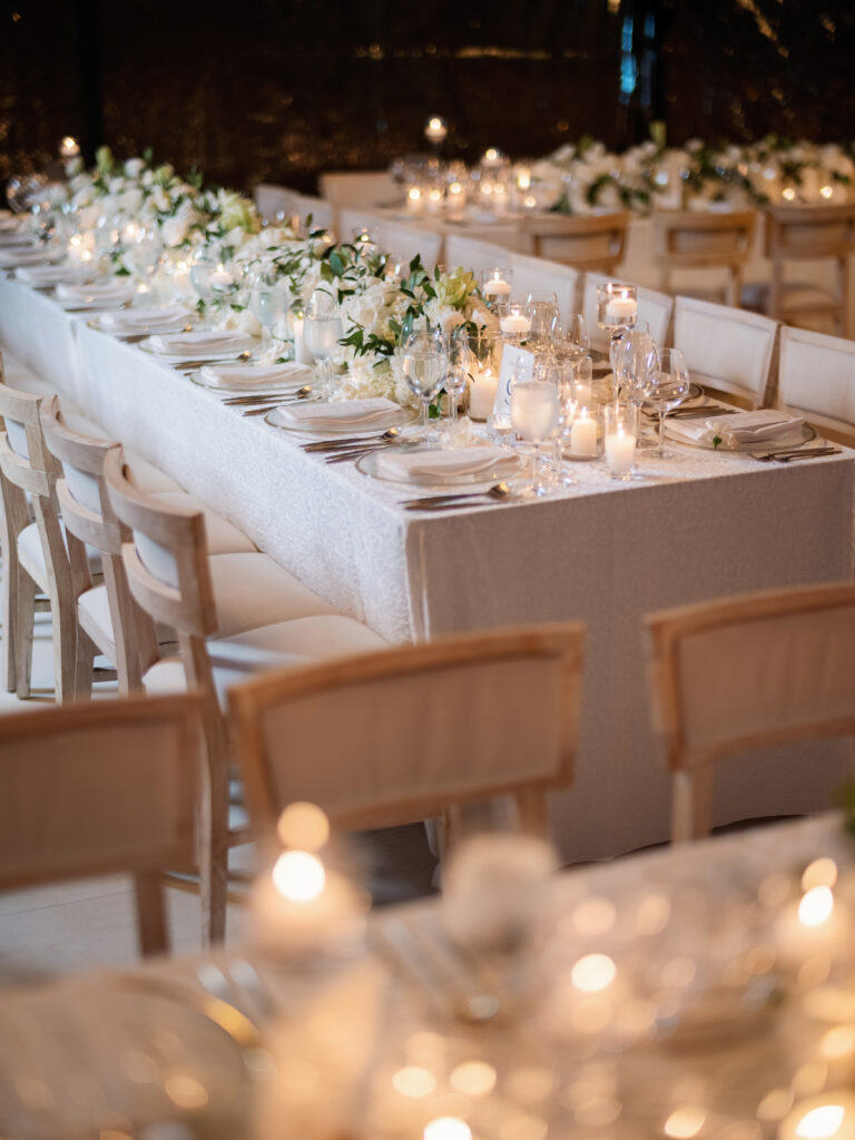 A long table set for a formal dinner event, featuring white tablecloth, elegant dishware, candles, and floral centerpieces. Rows of wooden chairs line both sides of the table.
