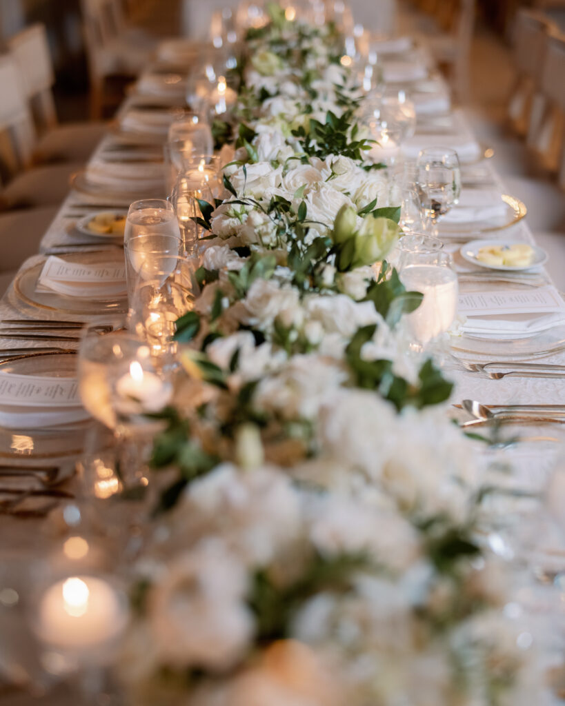 A long dining table with a centerpiece featuring white flowers and greenery, surrounded by glassware and white plates, is elegantly set for a formal event.