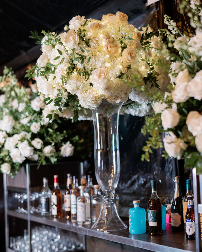 Large floral arrangement of white roses and greenery in a tall vase, placed on a bar counter with an assortment of liquor bottles and glassware.