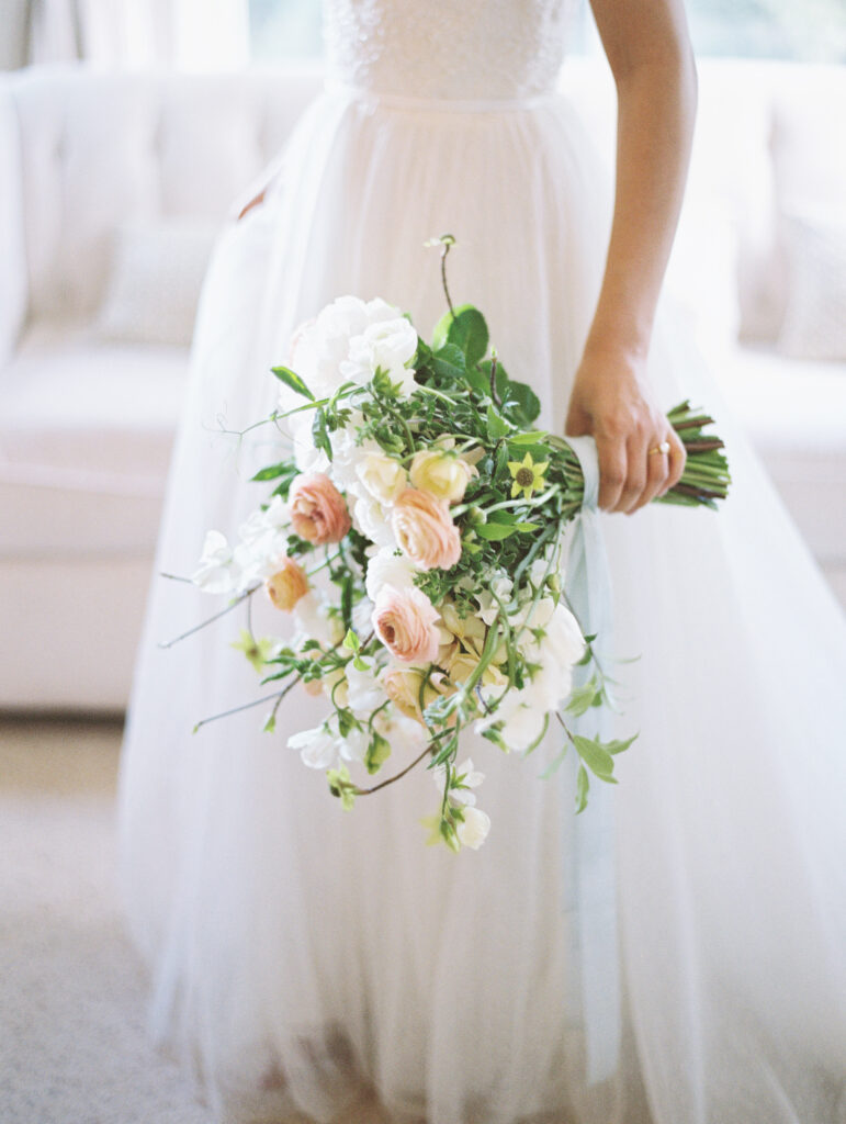 A bride in a white dress holds a bouquet of white and pink flowers with greenery, standing in a softly lit room.