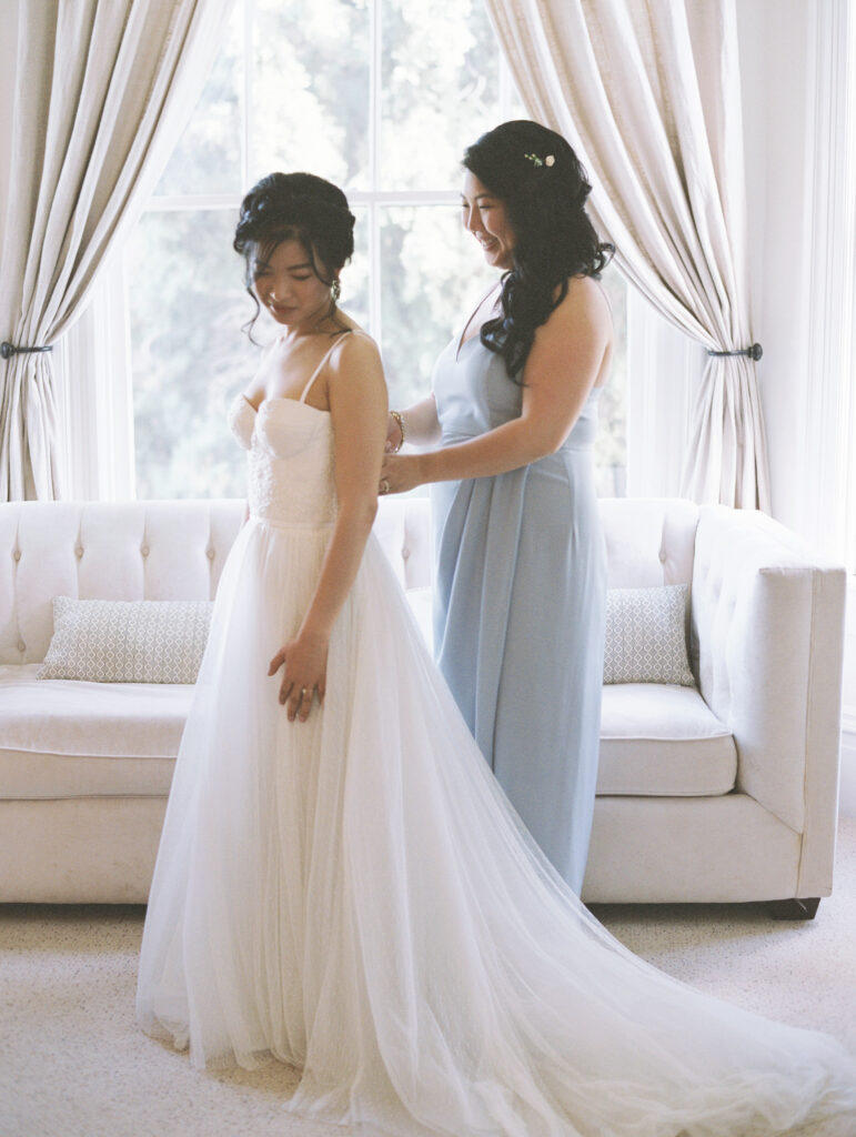 A bride in a white gown stands while another woman in a light blue dress adjusts her dress in front of a sofa by a window.