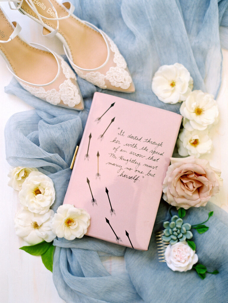 Flat lay of a pink book with a quote, surrounded by white and blush flowers, a light blue cloth, and white lace high-heeled shoes.