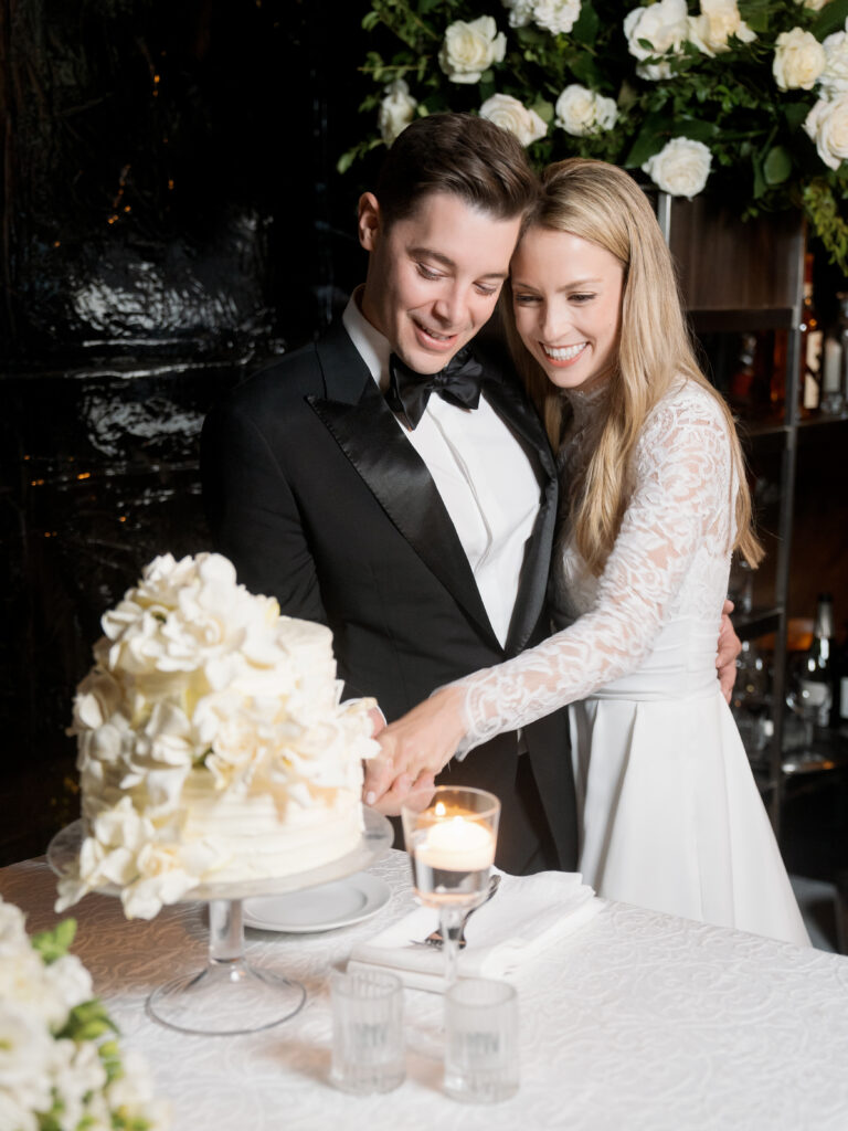 A bride and groom in formal attire cut a white tiered cake adorned with flowers. They are smiling and standing close together.