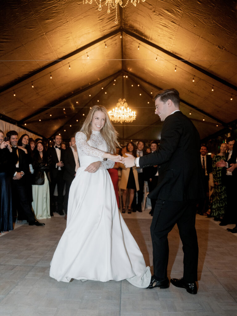 A couple dances at a formal event under a chandeliers and strings of lights, surrounded by a crowd of elegantly dressed onlookers.