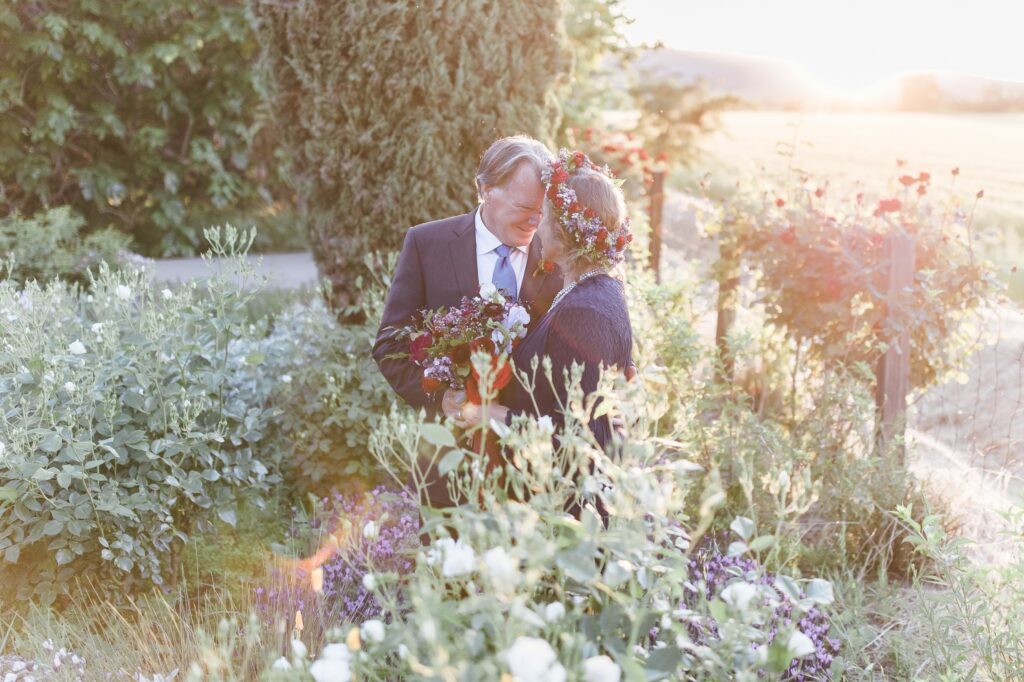 A couple embraces in a garden filled with lush greenery and flowers at sunset. The man wears a suit and the woman has a floral wreath on her head and holds a bouquet.