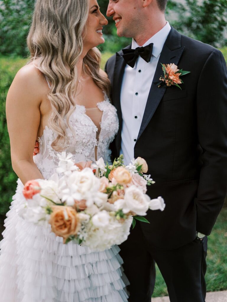 A bride and groom stand smiling at each other. The bride holds a bouquet of white and peach flowers. The groom wears a black tuxedo with a floral boutonniere.