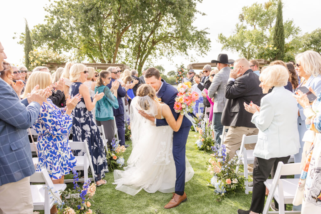 A newlywed couple kisses at the end of an outdoor aisle. Guests stand and applaud. The scene is decorated with flowers.