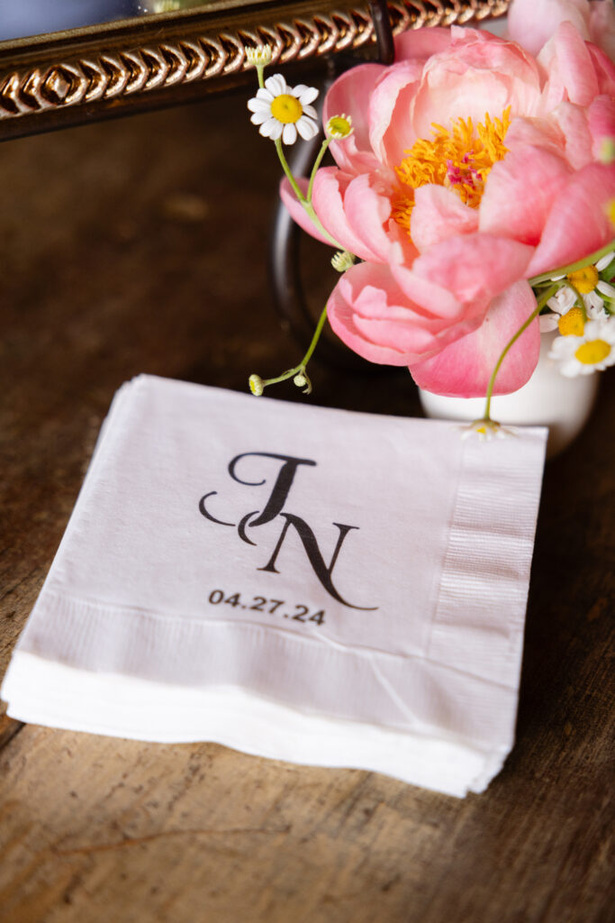A stack of personalized napkins with the initials "J N" and the date "04.27.24" printed on them, next to a pink flower on a wooden surface.