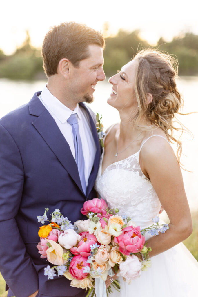 A couple in formal attire smiles at each other outdoors. The woman holds a bouquet of colorful flowers.