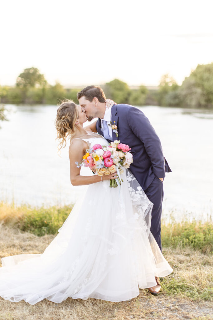 A couple in wedding attire kisses by a riverside. The bride holds a colorful bouquet.