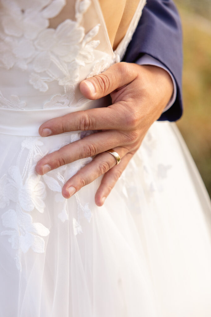A close-up of a hand wearing a wedding band resting on the back of a person in a white floral dress.