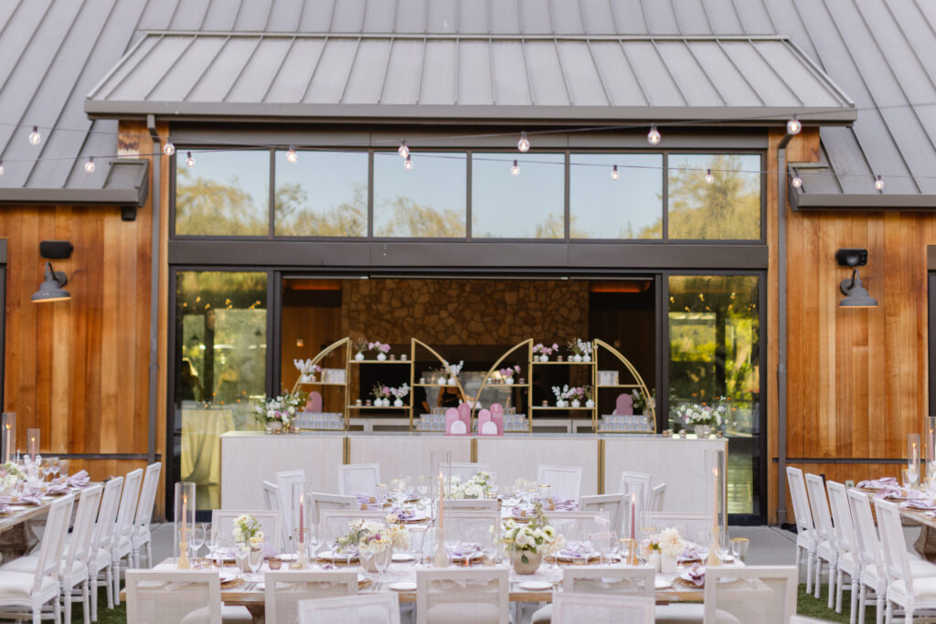 Outdoor wedding reception setup with tables decorated with white chairs, floral centerpieces, and string lights. A large wooden building is in the background.