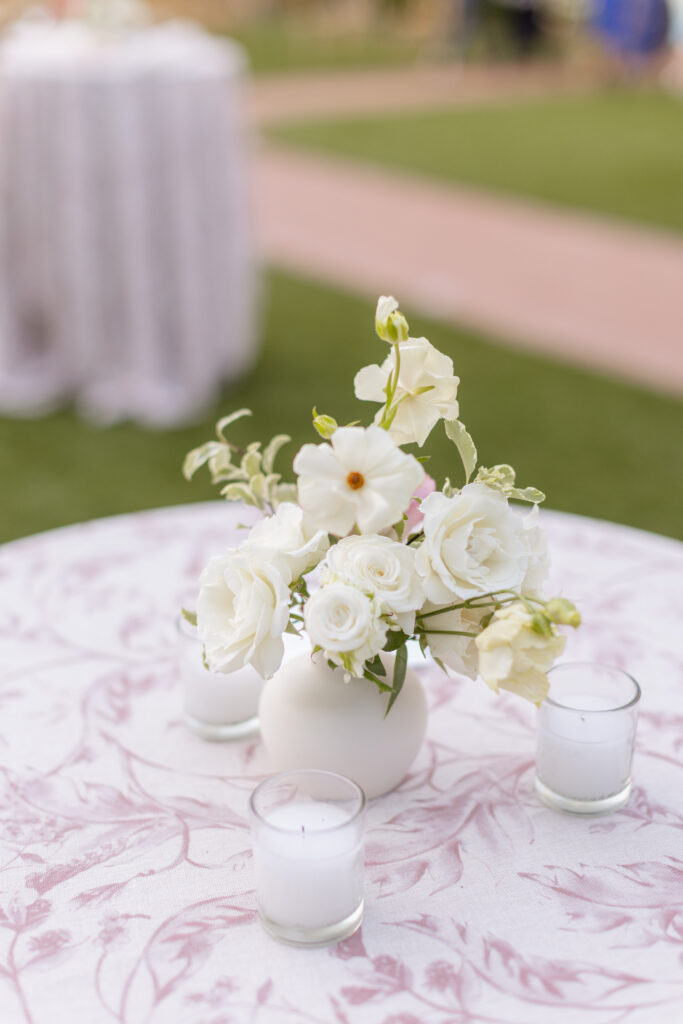 A white floral arrangement in a round vase is centered on a patterned tablecloth, surrounded by three small glass candles on a grassy outdoor setting.