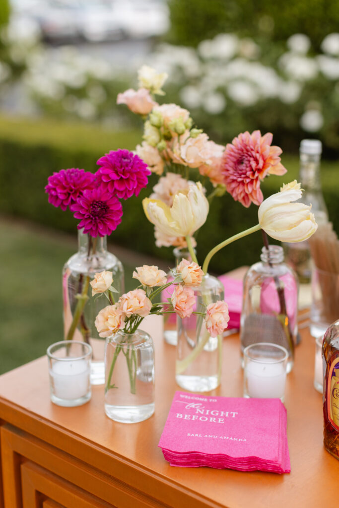 Glass vases with assorted flowers and small candles are arranged on a wooden table with bright pink napkins that read “The Night Before,” evoking the thoughtful touch of an Estate Yountville Wedding Planner.