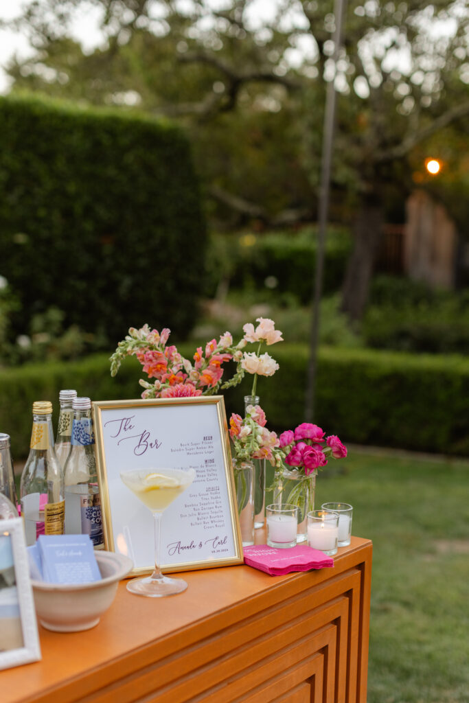 An outdoor bar setup curated by an expert Estate Yountville Wedding Planner features bottles, a cocktail menu in a gold frame, fresh flowers, and candles atop an orange table, beautifully arranged in a garden setting.