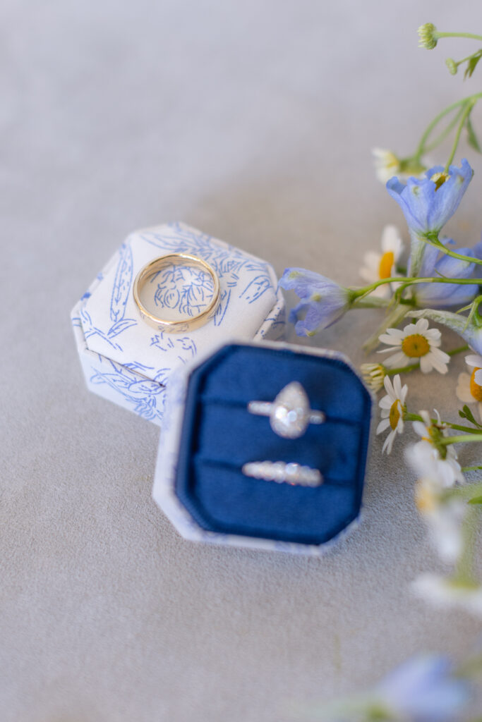 Two rings in decorative boxes: a gold ring in a floral box and a pear-shaped diamond ring with band in a navy blue box, next to small blue and white flowers.