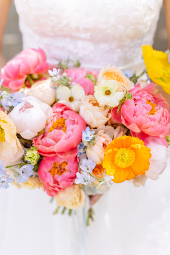 A person in a white dress holds a colorful bouquet with pink, yellow, orange, and white flowers, including peonies and poppies.