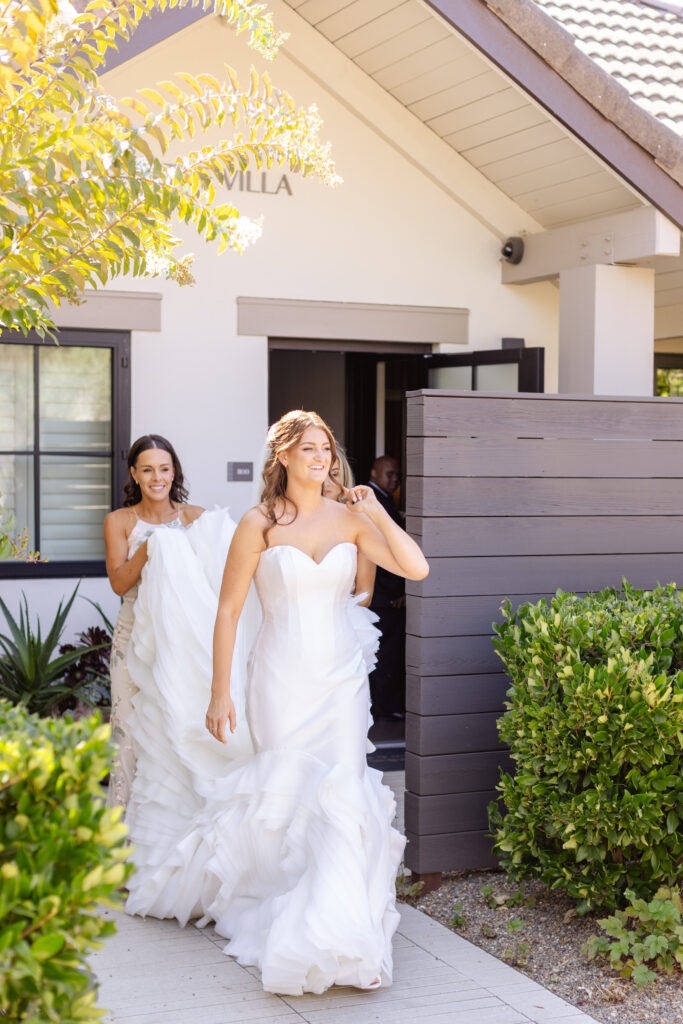 A bride in a white gown walks outside a house, smiling, while another woman helps with her train. Green bushes and a wooden fence line the walkway, beautifully arranged by an Estate Yountville Wedding Planner.