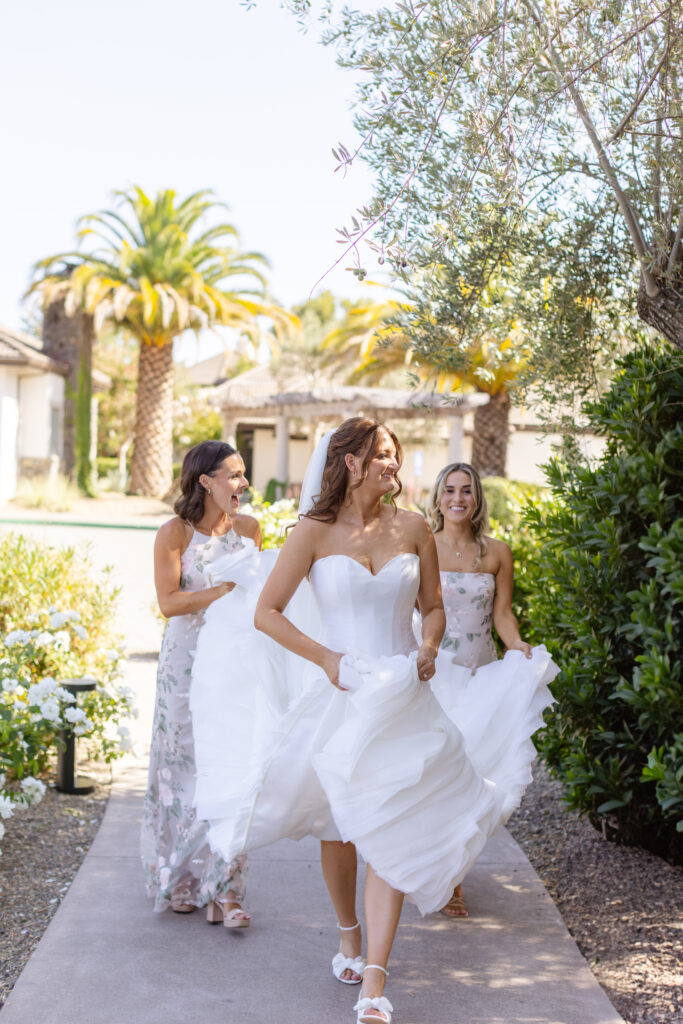 A bride in a white dress walks outside on a path, smiling, with two bridesmaids in floral dresses following behind her on a sunny day—every detail perfectly orchestrated by the Estate Yountville Wedding Planner.