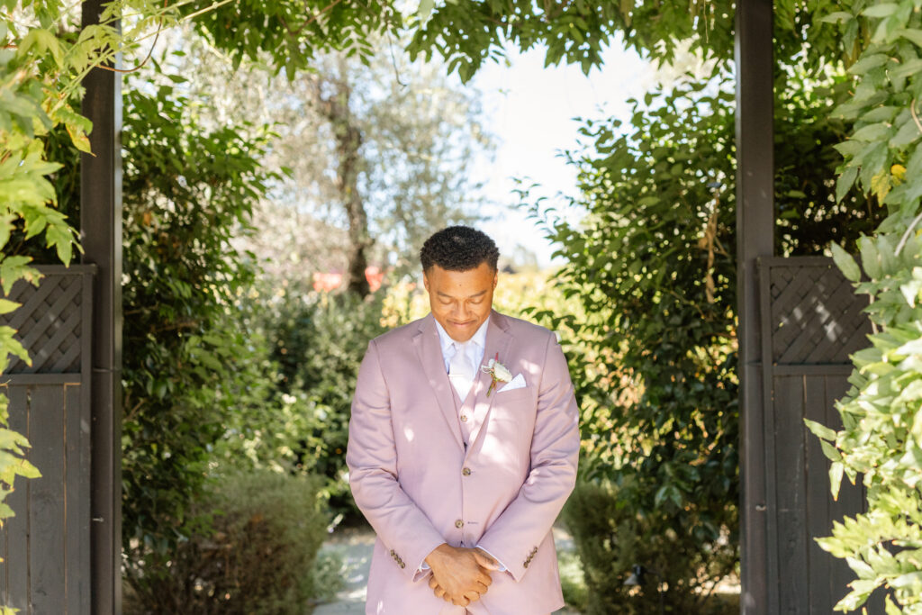 A man in a light purple suit stands under a leafy archway outdoors, thoughtfully looking down with his hands clasped in front of him—a serene moment beautifully coordinated by Estate Yountville Wedding Planner.