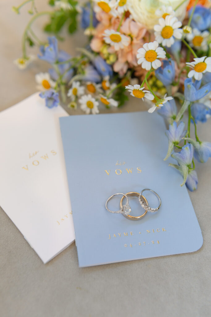 Three wedding rings rest on a blue vows booklet labeled "his Vows," with flowers and a white vows booklet nearby.