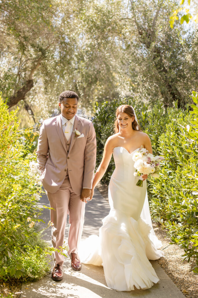 Bride in a white strapless gown and groom in a light tan suit walk hand in hand outdoors, surrounded by greenery and sunlight, beautifully orchestrated by an expert Estate Yountville Wedding Planner.