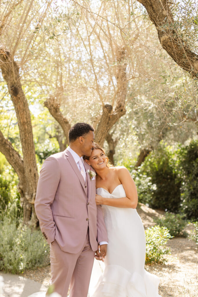 A bride in a white dress leans on a groom in a mauve suit as they stand outdoors under sunlit trees, both smiling—a picture-perfect moment beautifully orchestrated by an Estate Yountville Wedding Planner.