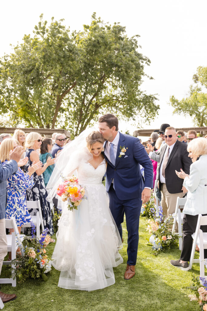 A bride and groom walk down a grassy aisle outdoors, surrounded by guests applauding. The bride holds a colorful bouquet, and both are smiling.